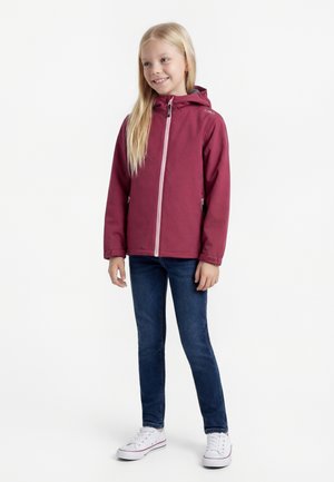 Dievča s dlhými blond vlasmi, oblečené v bordovej kapucňovej bunde, tmavomodrých džínsoch a bielych teniskách, stojace a usmievajúce sa na bielom pozadí.