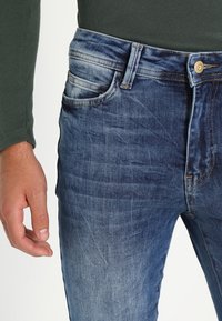 Tummansiniset farkut, joissa on haalistunut pinta. Ominaisuuksiin kuuluu viisi taskua, napitus ja suora lahjetyyli. Rakennehenkilöitys.