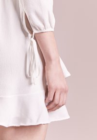 Bílá strukturovaná šaty s nabíranými rukávy, se zavazováním na manžetách a volánkovým lemem. Detailní záběr na ruku držící látku.