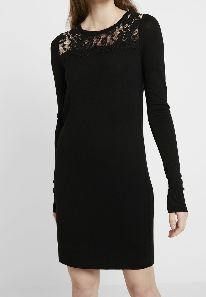 Femme portant une robe noire à manches longues avec un panneau en dentelle fleurie sur la partie supérieure de la poitrine et les épaules.