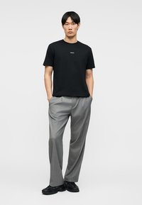 Άνδρας όρθιος με τα χέρια στις τσέπες, φοράει μαύρο μπλουζάκι με μικρό λευκό κείμενο, γκρι παντελόνι και μαύρα παπούτσια σε απλό φόντο.