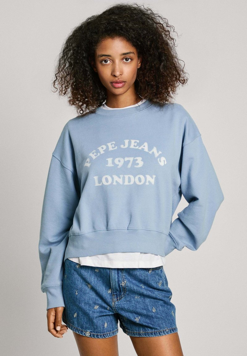Lichtblauwe oversized sweatshirt met witte opdruk "PEPE JEANS 1973 LONDEN". Heeft een ronde halslijn en geribbelde zoom. Gecombineerd met denim shorts.