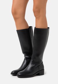 Bottes noires montant jusqu'au genou, fabriquées en cuir synthétique lisse, avec un bout arrondi, un petit talon carré et une fermeture éclair sur le côté pour un enfilage facile.