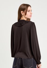 Blouse noire avec des manches longues bouffantes, un col à volants et des poignets froncés. Fabriquée en tissu doux avec une texture lisse.