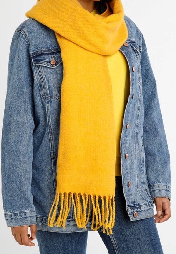 Personne portant une écharpe jaune vif à franges sur un haut jaune et une veste en jean bleue avec un jean.