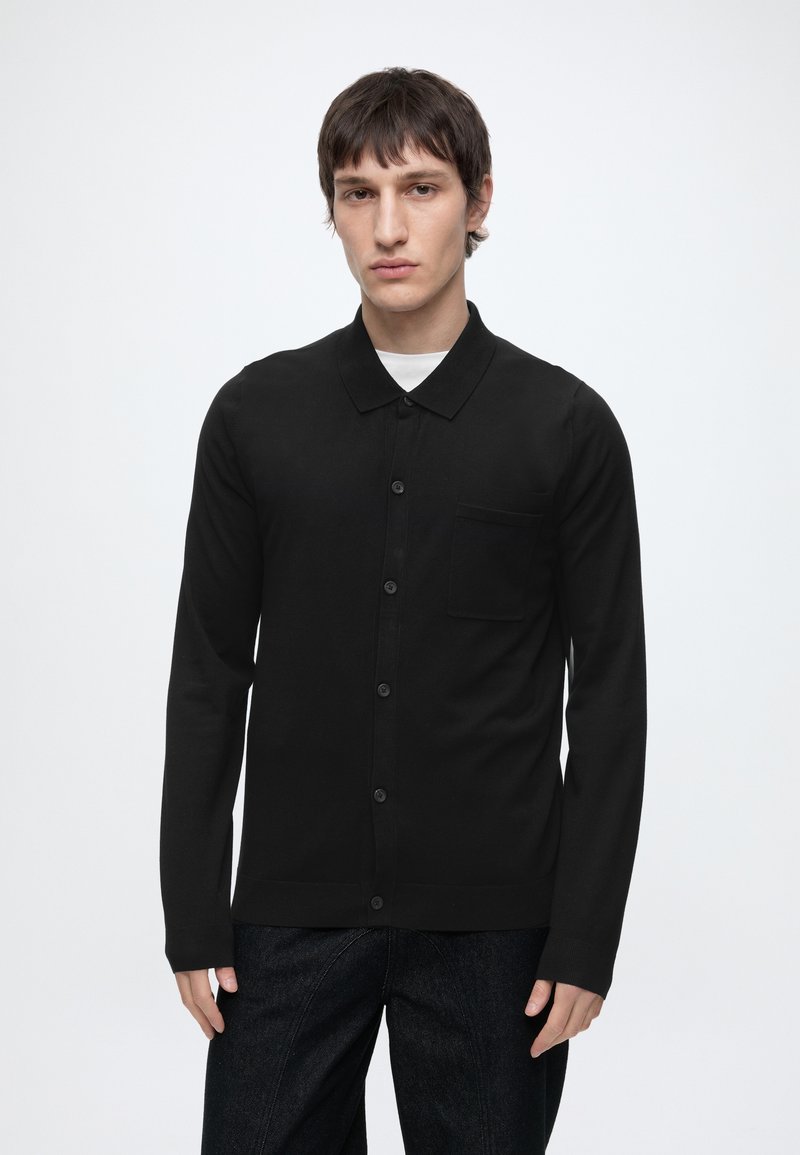 Jeune homme aux cheveux bruns courts portant une chemise noire à manches longues boutonnée par-dessus un sous-vêtement blanc, sur un fond clair uni.