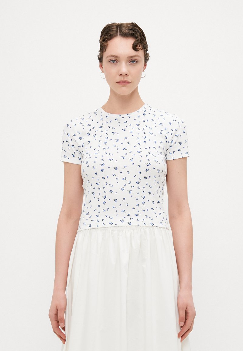 Wit T-shirt met korte mouwen en blauw bloemmotief, ronde halslijn, aansluitende body en een zachte textuur. Gecombineerd met een vloeiende witte rok.