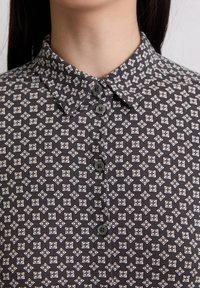 Camisa de rayas en blanco y negro con un cuello con solapa y tres botones grises. Presenta un diseño geométrico con pequeños acentos florales.