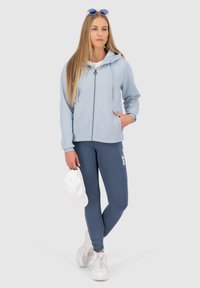 Felpa con zip azzurro chiaro con coulisse, abbinata a leggins navy con testo bianco. Il modello tiene un cappellino bianco. Sneakers bianche completano l'outfit.