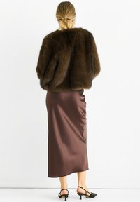 Woman with blonde hair in low ponytail wearing a dark brown fur jacket, satin midi skirt, and black heeled slingback shoes, standing with back to camera.