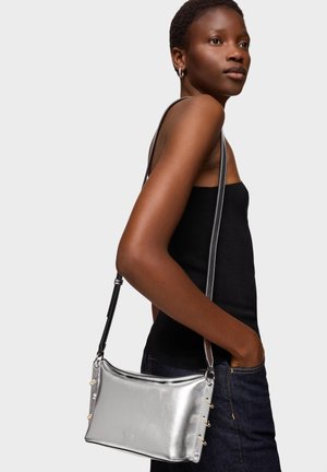 Kobieta o krótkich włosach, ubrana w czarny, bezramienny top i ciemne dżinsy, nosząca metaliczną srebrną torebkę na ramię z bocznymi sznurowaniami.