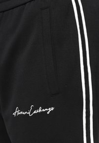 Svarte joggebukser i mykt stoff, med hvite sidestriper og et brodert logo på frontlommens område.
