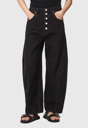 Person iført løstsiddende sorte jeans med fem forreste knapper og sorte sandaler, stående med hænderne i lommerne mod en ensfarvet baggrund.