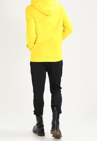 Žlutá mikina s kapucí a klokáním kapsou, doplněná černými zúženými tepláky. Černé kotníkové boty se zipem vzadu.