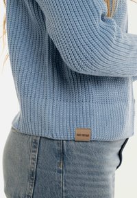 Lichtblauwe gebreide trui met een geribbelde structuur, met een klein bruin label aan de zoom met de tekst "KNIT FACTORY." Draagt met denim jeans.