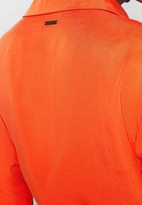 Ljus orange skjorta med en slät, lätt glänsande textur. Har en spetsig krage och en liten märkesetikett nära kragen på baksidan.