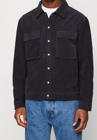 Sametová bunda v tmavě námořnické barvě s dvěma náprsními kapsami, zapínáním na knoflíky a texturovaným povrchem. Spárována s světle modrými džíny.