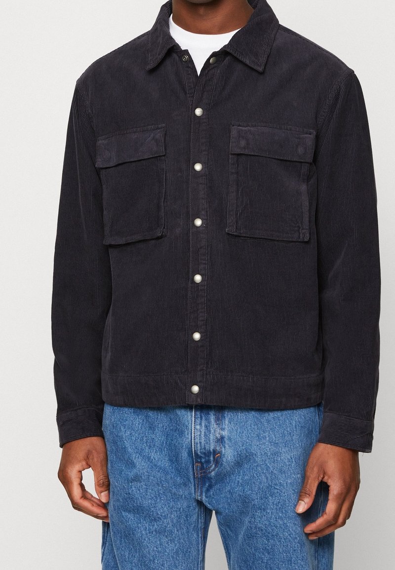 Sametová bunda v tmavě námořnické barvě s dvěma náprsními kapsami, zapínáním na knoflíky a texturovaným povrchem. Spárována s světle modrými džíny.