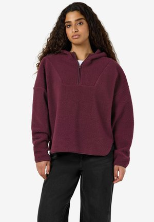 Femme aux longs cheveux bouclés portant un pull polaire bordeaux avec une courte fermeture éclair et un pantalon noir, debout devant un fond uni.
