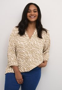Lächelnde Frau mit langen dunklen Haaren, die eine beige und weiße, zebra-gestreifte Bluse und blaue Jeans trägt, steht vor einer hellen Wand.