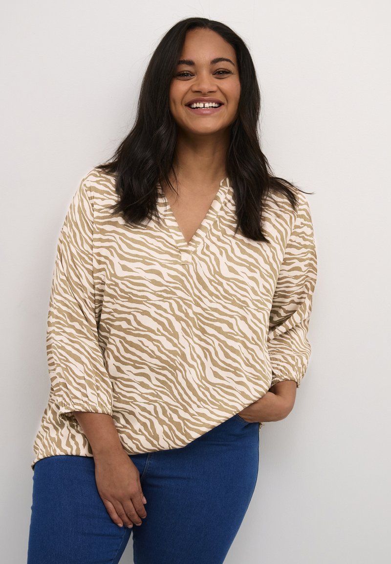 Smilende kvinde med langt mørkt hår iført en bluse med beige og hvid zebra-stribe og blå jeans, stående mod en lys væg.