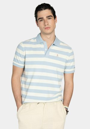 Joven con cabello oscuro que lleva una camisa tipo polo a rayas azul claro y blanca y pantalones beige, de pie con las manos en los bolsillos.