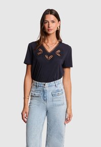 Camiseta de color azul marino con un escote en V decorativo que presenta recortes en forma de corazón, complementada con mangas cortas y combinada con unos vaqueros de tiro alto en denim claro.