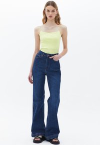 Top fitted giallo chiaro con spalline sottili, abbinato a jeans blu scuro a vita alta che si allargano nella parte finale, indossato con sandali neri.