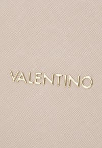 Țesătură texturată de culoare roz deschis, prezentând numele brandului "VALENTINO" în litere aurii embosate. Design curat și modern, cu detalii subtile.