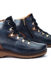Botas de tobillo de cuero azul oscuro con detalles en marrón, herrajes dorados y una suela texturizada. Presenta cordones y un cierre con cremallera para facilitar el uso.
