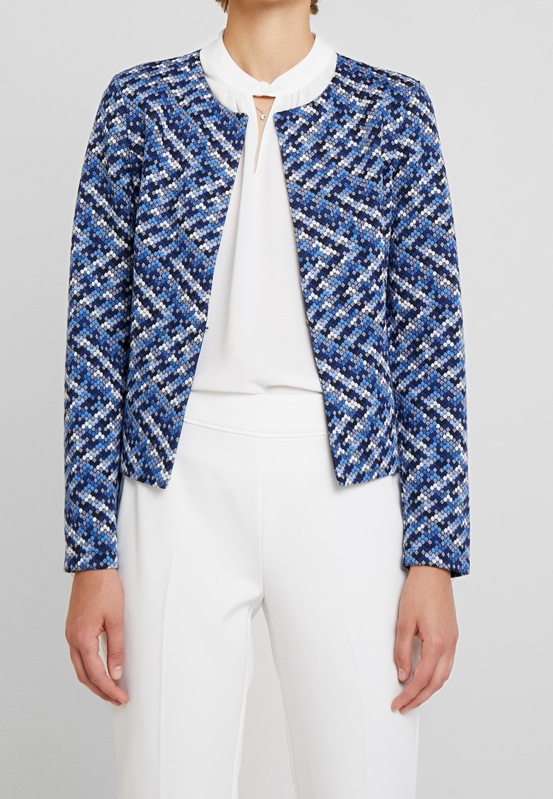 Femme portant une veste à manches longues bleue à motifs par-dessus un chemisier blanc et un pantalon blanc, debout devant un fond gris uni.