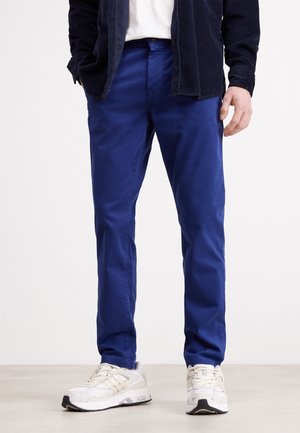 Pantalon en coton bleu avec une coupe ajustée, texture lisse et poches latérales, associé à des chaussures de sport blanches. Design simple, sans motifs marquants.
