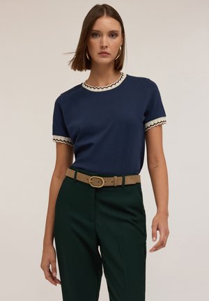 Camiseta de manga corta azul marino con un escote y mangas en crema con forma de ondas. Combinada con unos pantalones verdes de cintura alta y un cinturón beige con una hebilla dorada.