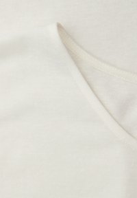 Zachte witte stof met een close-up van overlappende afgeronde halsnaadnaden, waarbij fijne stikdetails en een gladde textuur zichtbaar zijn.