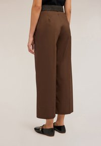 Pantalons larges marron avec une ceinture élastique noire texturée, présentant une finition lisse et un ourlet court, associés à des chaussures noires sans lacets.