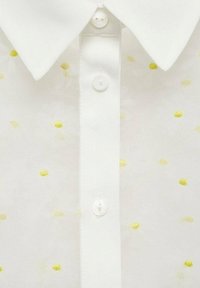 Camicia bianca con colletto classico, caratterizzata da sottili motivi floreali e piccoli accenti gialli. I bottoni sono circolari e trasparenti.