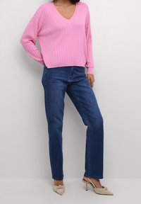 Pull en maille côtelée rose avec col en V, associé à un jean bleu marine à jambes droites et des escarpins beiges  à bout pointu avec un détail noué.