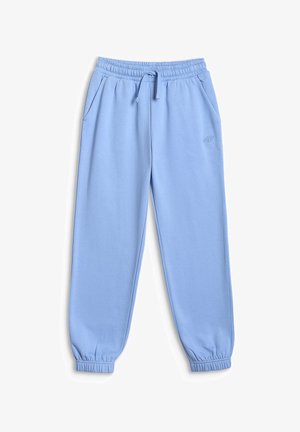 Jeune fille aux longs cheveux bruns portant une veste crème et un haut, un pantalon de survêtement bleu clair, des baskets blanches, tenant une pochette noire dans la main droite.