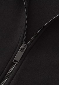 Dettaglio di un indumento nero con zip, caratterizzato da una finitura opaca liscia e una zip robusta con un tirante rettangolare, su uno sfondo tessile.