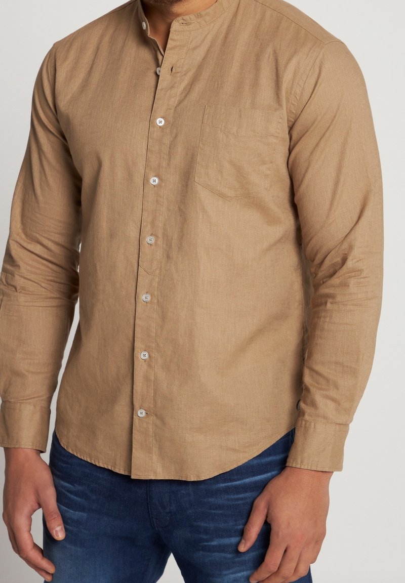 Homme portant une chemise beige à manches longues boutonnée avec un col mandarin et un jean bleu sur un fond uni.