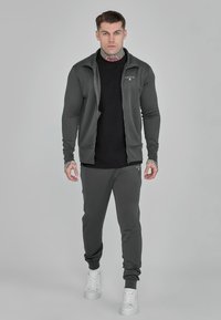 Dark gray athletic tracksuit with a zip-up jacket and slim-fit pants. Fabric is smooth and stretchy. Features a small logo on the jacket.