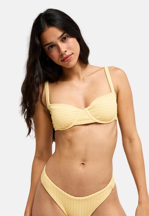 Young woman with long dark hair wearing a textured light yellow bikini with underwire top against a plain white background.
