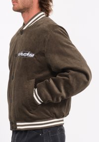 Bruine corduroy bomberjack met witte gestreepte manchetten en zoom, voorzien van een knoopsluiting en een opvallend logo op de borstzak.