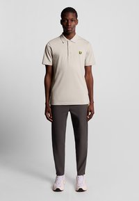 Beige polo shirt met een kraag en korte mouwen, met een klein geel logo. Gepaard met donkergrijze broek en lichte sneakers.