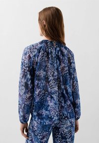 Long-sleeved blouse in sheer blue fabric with abstract purple and black patterns. Features gathered neckline and button detail at the back.