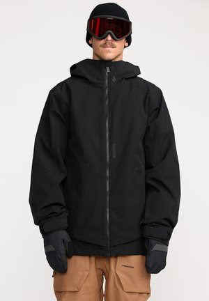 Veste de neige noire avec fermeture éclair à l'avant, capuche ajustable, accompagnée de gants noirs. Le modèle porte un bonnet et des lunettes de protection teintées de rouge.