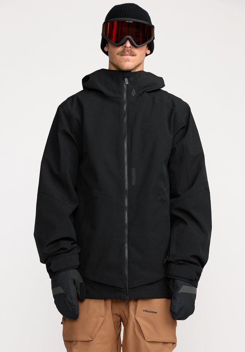 Veste de neige noire avec fermeture éclair à l'avant, capuche ajustable, accompagnée de gants noirs. Le modèle porte un bonnet et des lunettes de protection teintées de rouge.