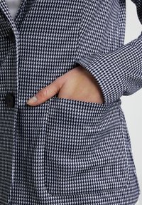 Veste à carreaux bleu marine et blanc, dotée d'un col cranté, d'une fermeture à boutons et d'une poche latérale avec une texture de tissu lisse.