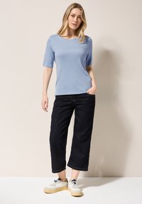 Světle modrý top s krátkým rukávem a jemně strukturovaným povrchem. Slučuje se s tmavými kalhotami se širokými nohavicemi a bílými teniskami se světlými podrážkami.