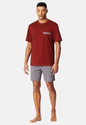 Rood T-shirt met korte mouwen en een blauwe geruite zak, gecombineerd met gepatineerde short in blauw en wit. Model is blootsvoets.
